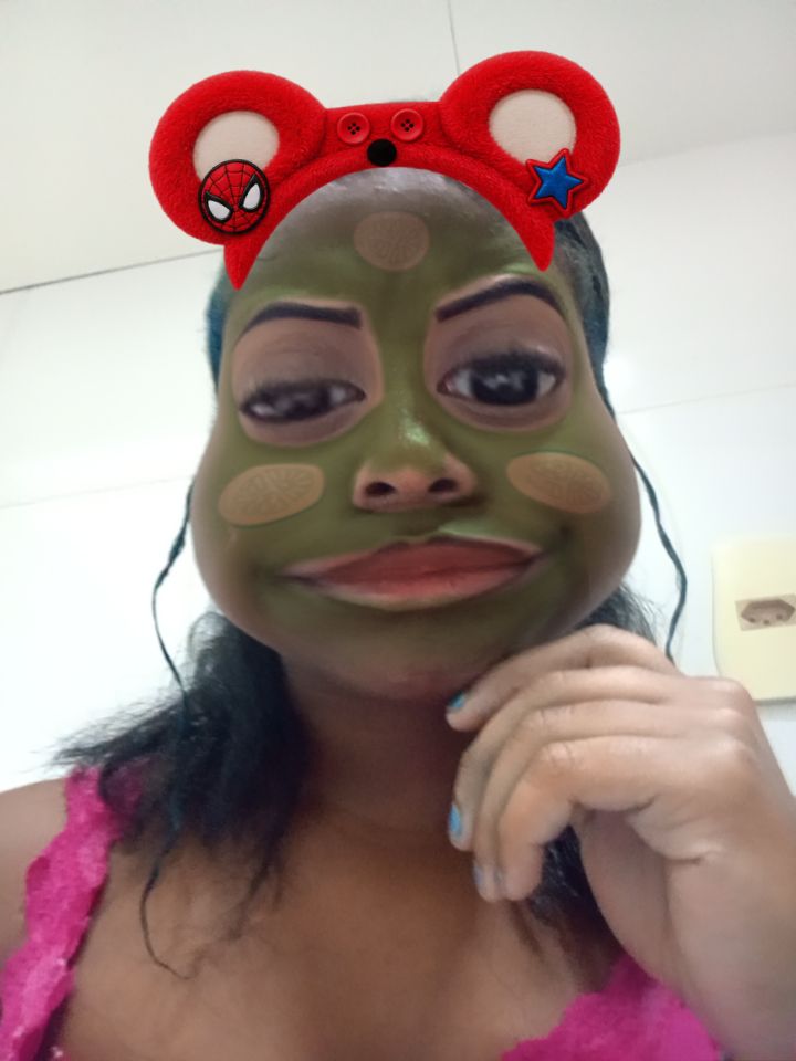 Mirela de máscara verde de sapo, tiara vermelha de orelhinhas, fazendo pose com a mão no queixo
